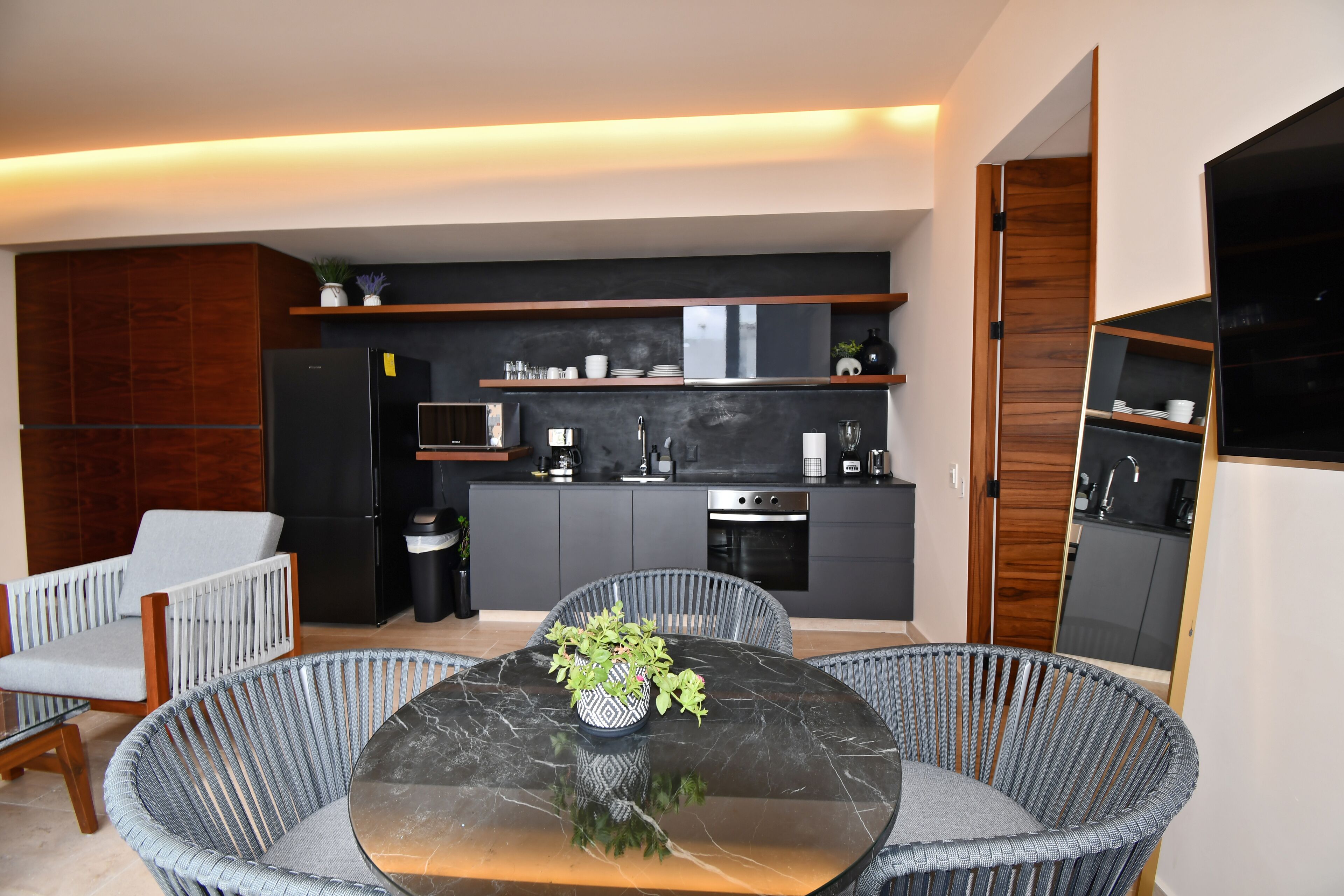 Een moderne keuken met donkere kastjes, een ingebouwde oven en een aanrecht met een spoelbak. Er is een eethoek met een glazen tafel en rieten stoelen.