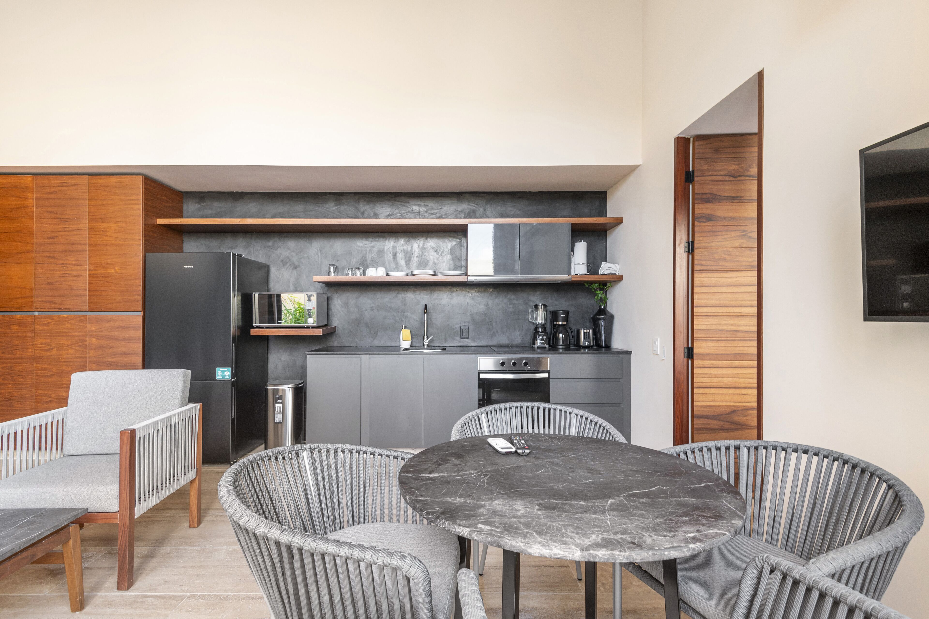 Uma cozinha moderna com armários cinza, geladeira preta e forno embutido. Há uma área de jantar com mesa redonda e cadeiras de vime.