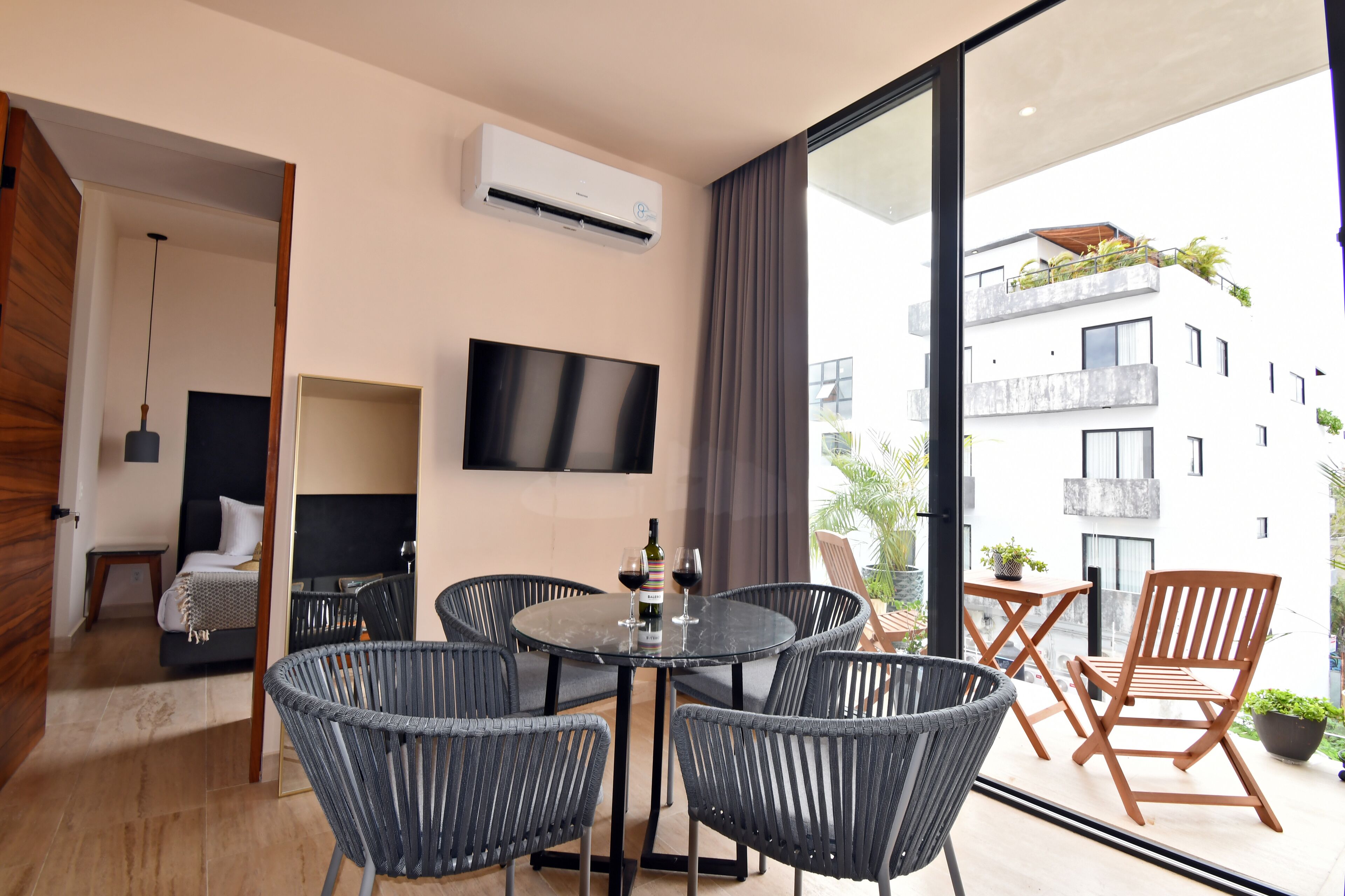 Una habitación moderna con una mesa redonda, cuatro sillas, un televisor de pantalla plana y un balcón con sillas de madera y plantas en macetas.