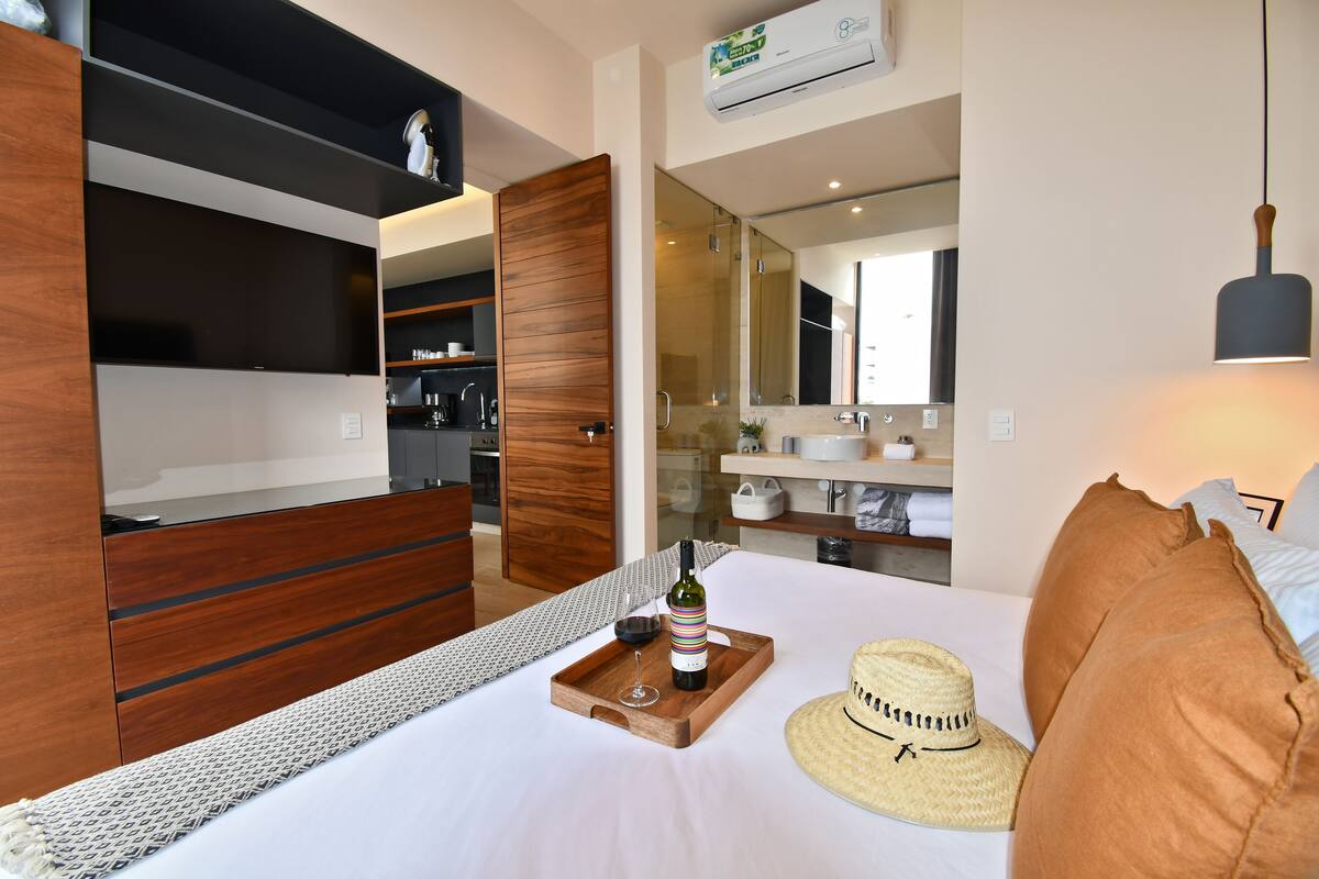 Una habitación de hotel moderna con un armario de madera, una mesa de comedor con una botella de vino y una copa, y un sombrero de paja en la cama.