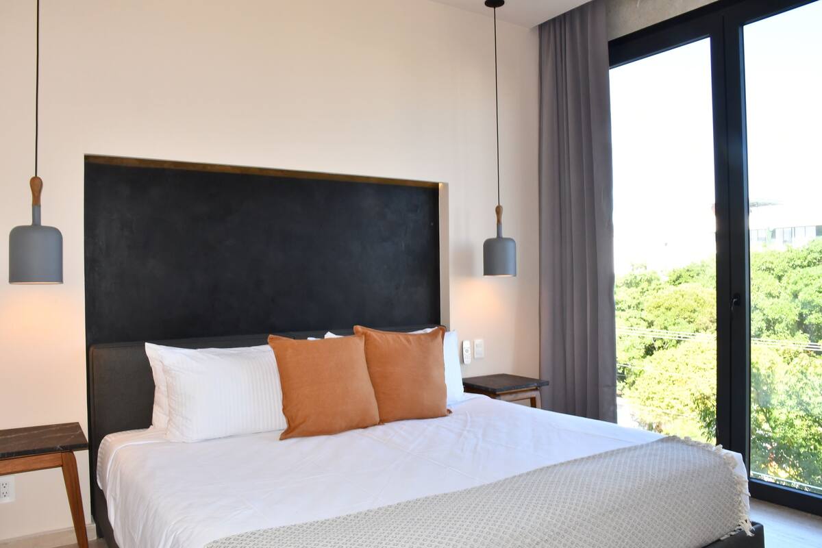 Una habitación de hotel moderna con una cama grande, dos mesitas de noche y vistas a zonas verdes por la ventana.