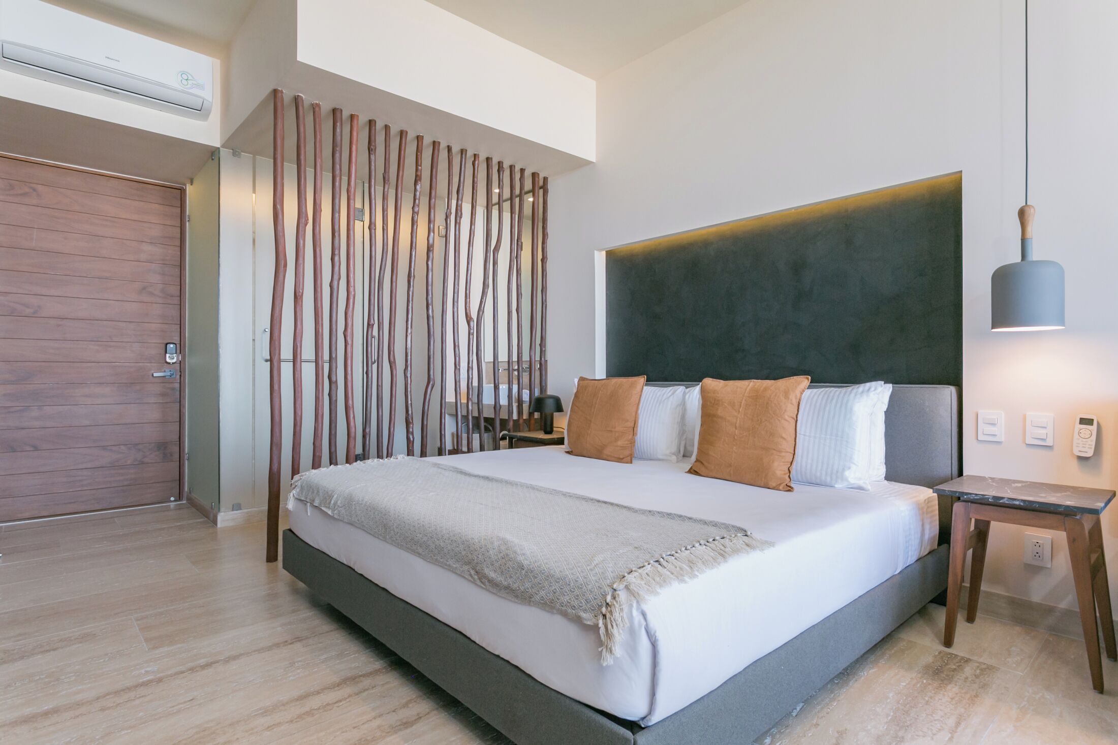 A modern bedroom with a large bed, a wooden slat partition, a bedside table with a lamp, and a wall-mounted air conditioner.