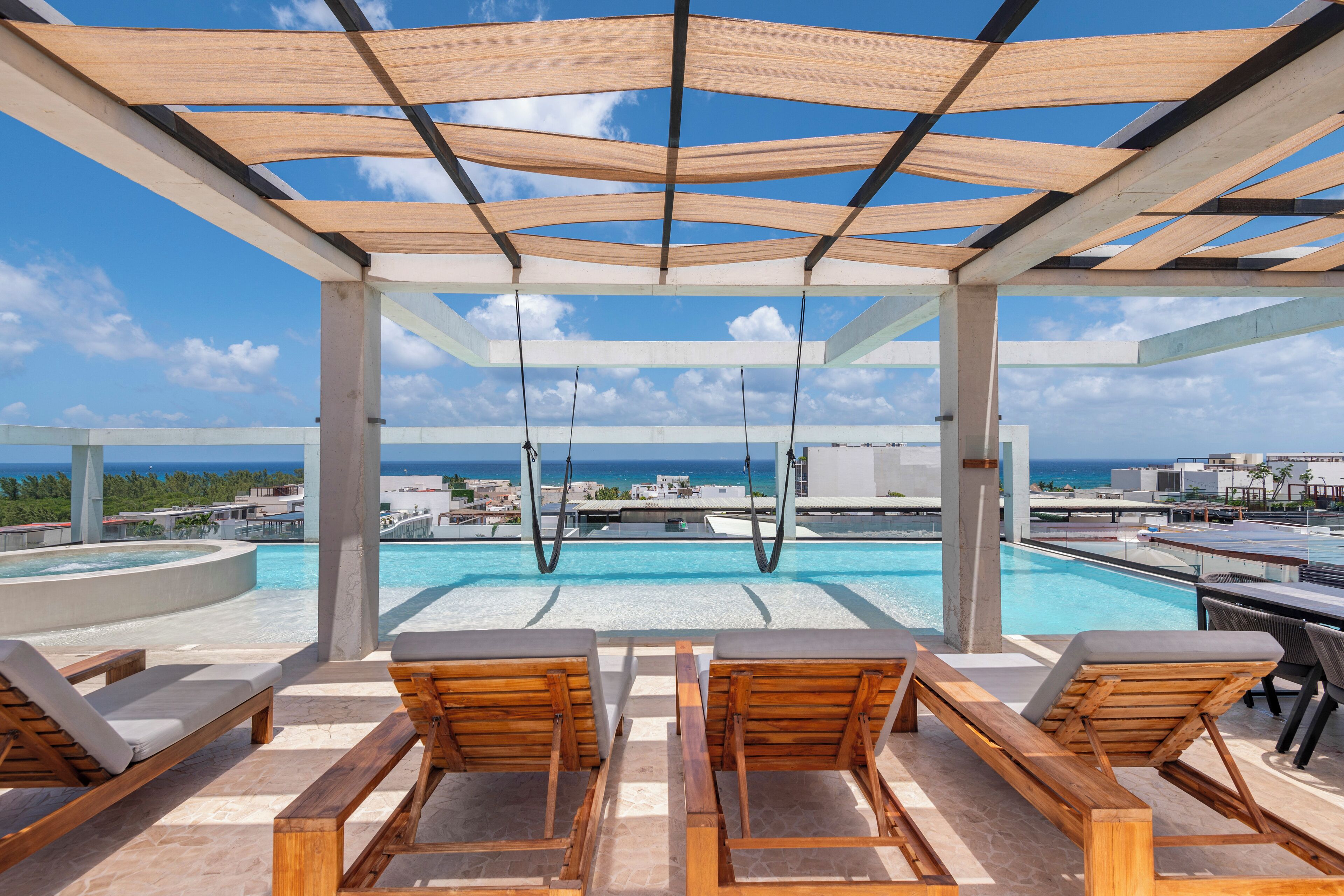 A rooftop pool area with wooden loungers, a swing, and a view of the ocean and cityscape.
