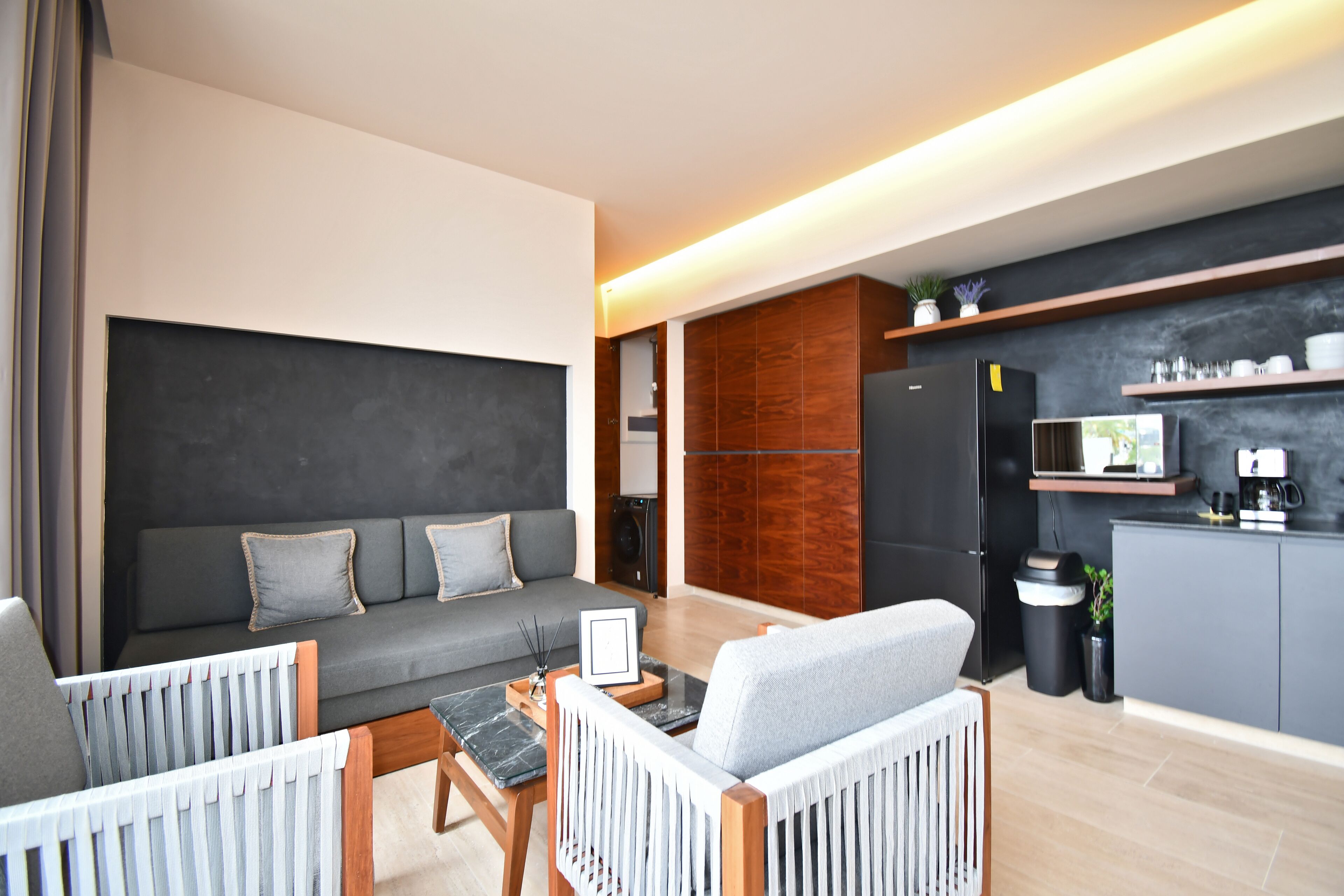 A modern living room with a grey sofa, wooden chairs, a coffee table, and a kitchen area with a microwave and cabinets.