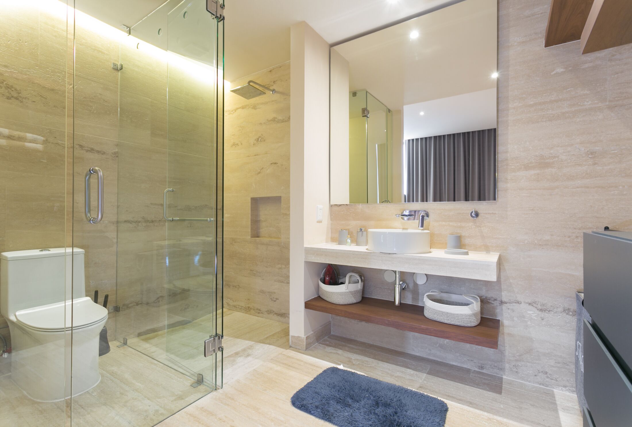 A modern bathroom with a glass-enclosed shower, a white toilet, a sink with a mirror above, and a blue rug on the floor.