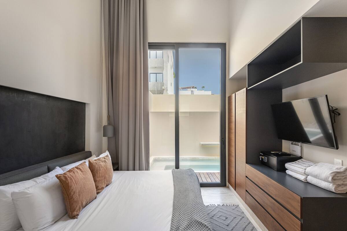 Una habitación de hotel moderna con una cama grande, un televisor, un escritorio con lámpara y vistas a una piscina y edificios.