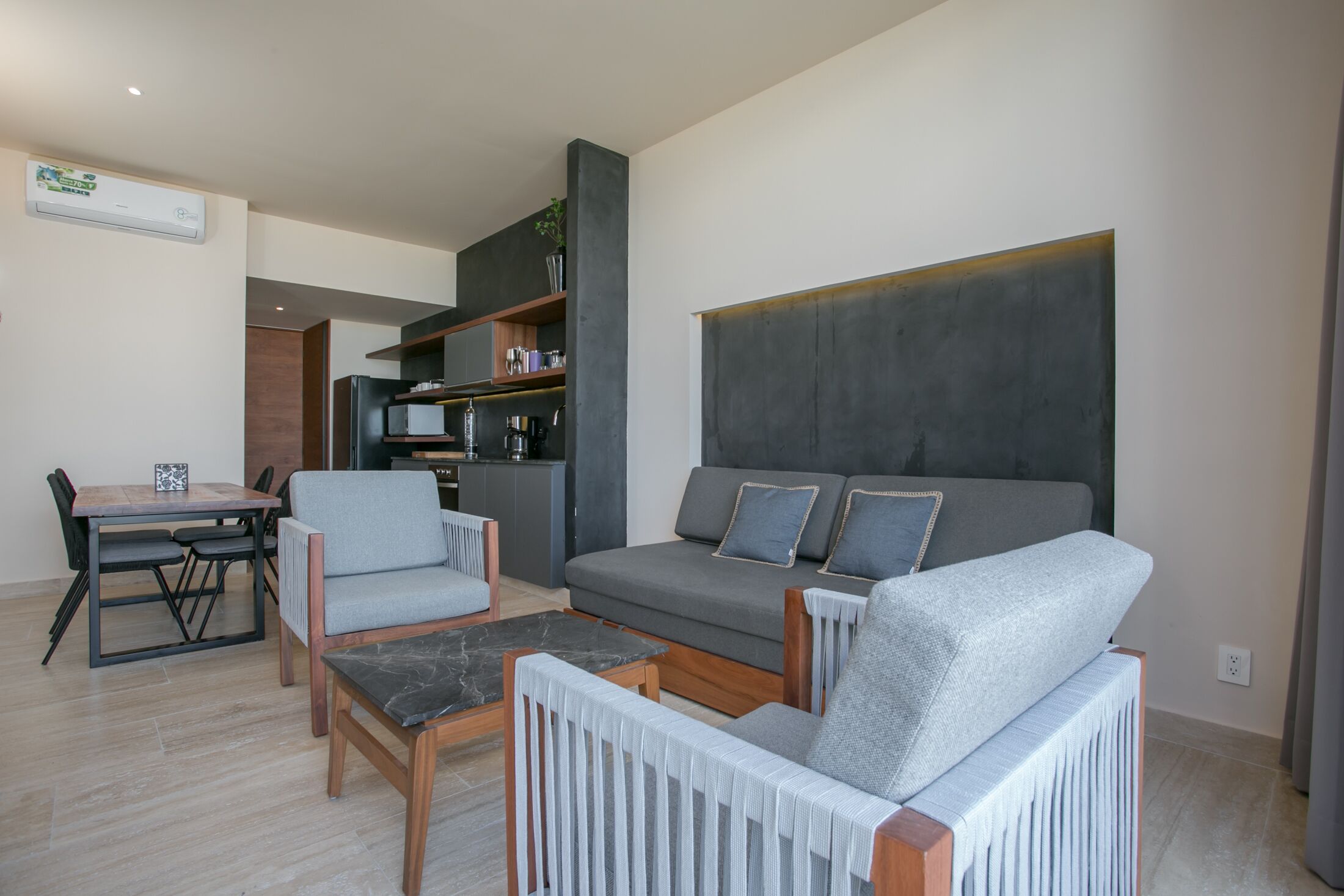 Een moderne woonkamer met een grijze bank, twee fauteuils, een salontafel en een eethoek met een houten tafel en stoelen.