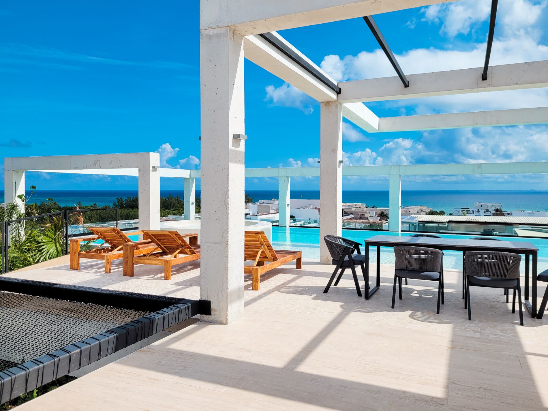 A rooftop terrace with a pool, wooden lounge chairs, and a table with chairs.