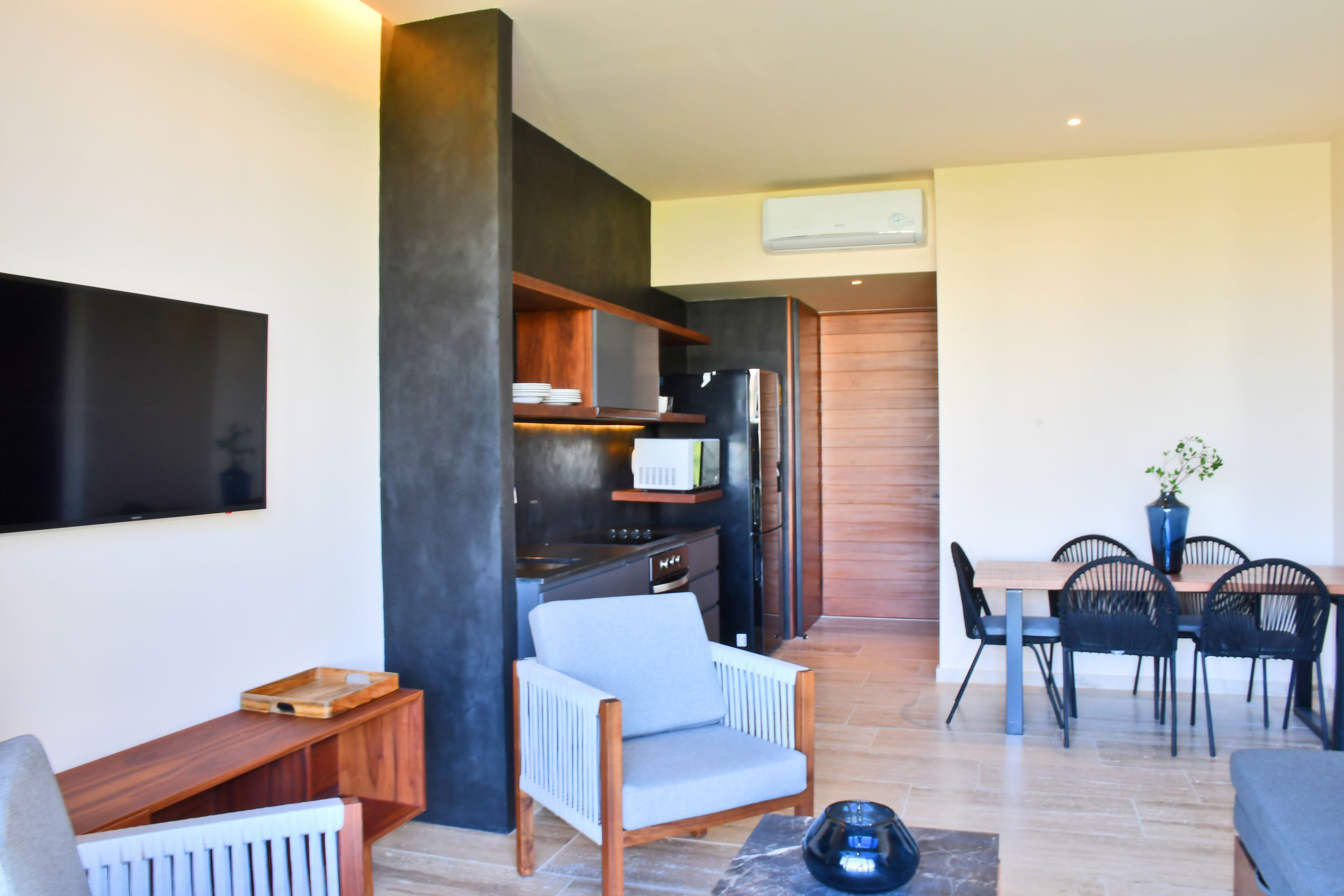 A modern living room with a sofa, a coffee table, a TV mounted on the wall, a dining area with a table and chairs, and a kitchen area with cabinets and appliances.