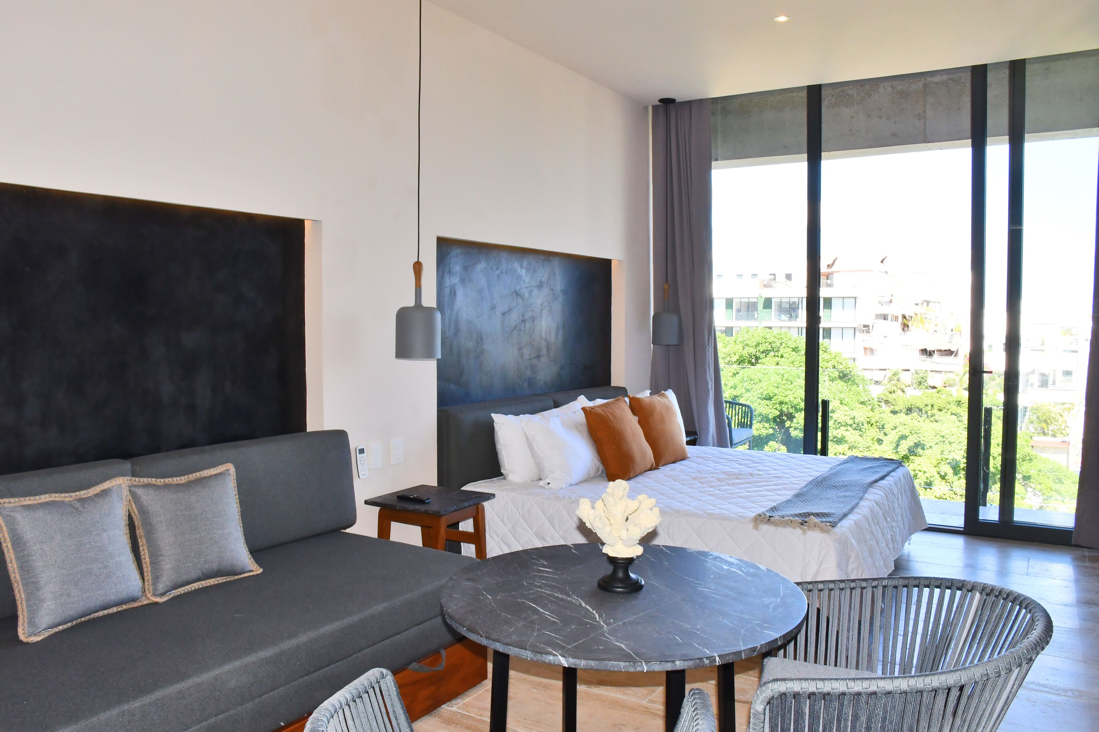 Una habitación de hotel moderna con una cama grande, un sofá, una mesa de comedor y vistas a la ciudad a través de amplios ventanales.