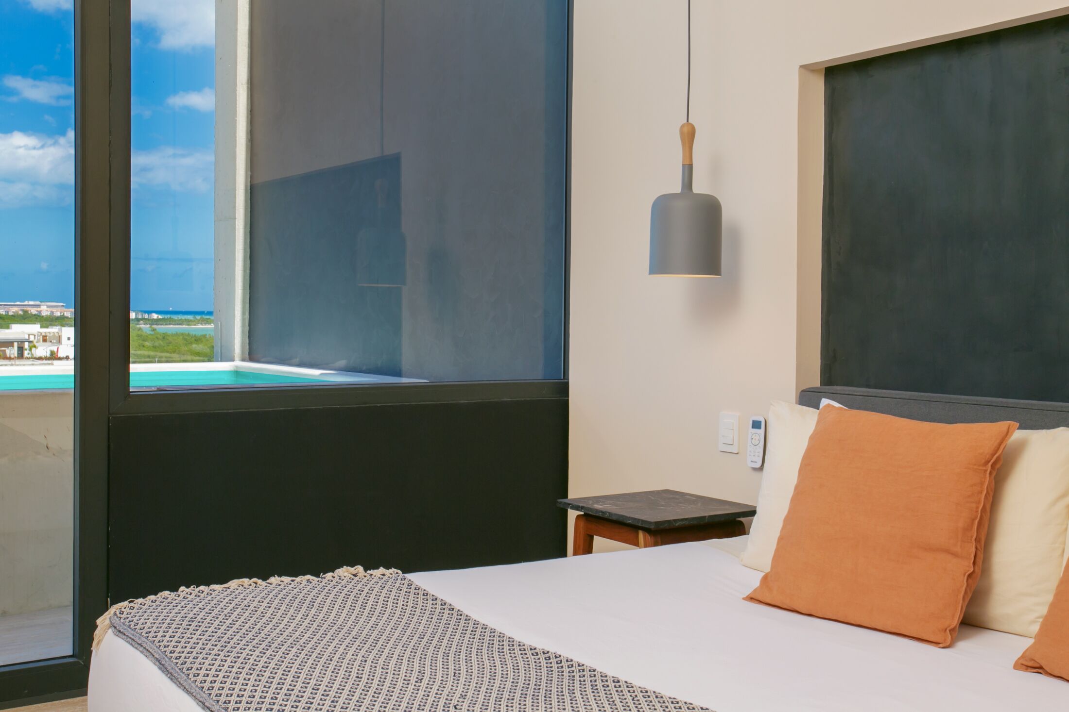 Habitación de hotel con un ventanal grande, una cama con dos almohadas, una mesita de noche y una lámpara colgante.