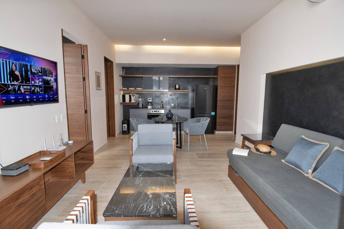 Una sala de estar moderna con sofá, mesa de centro y televisor. Se observa una cocina al fondo con una mesa de comedor y sillas.