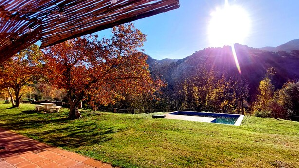 A pool area with a pergola, surrounded by grass and trees with autumn foliage, set against a mountainous backdrop. - Rural house, ideal for families in the middle of nature. (Júzcar)