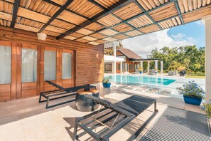 Un espacio exterior techado con piscina, sillas de descanso y un edificio de madera con amplias ventanas.