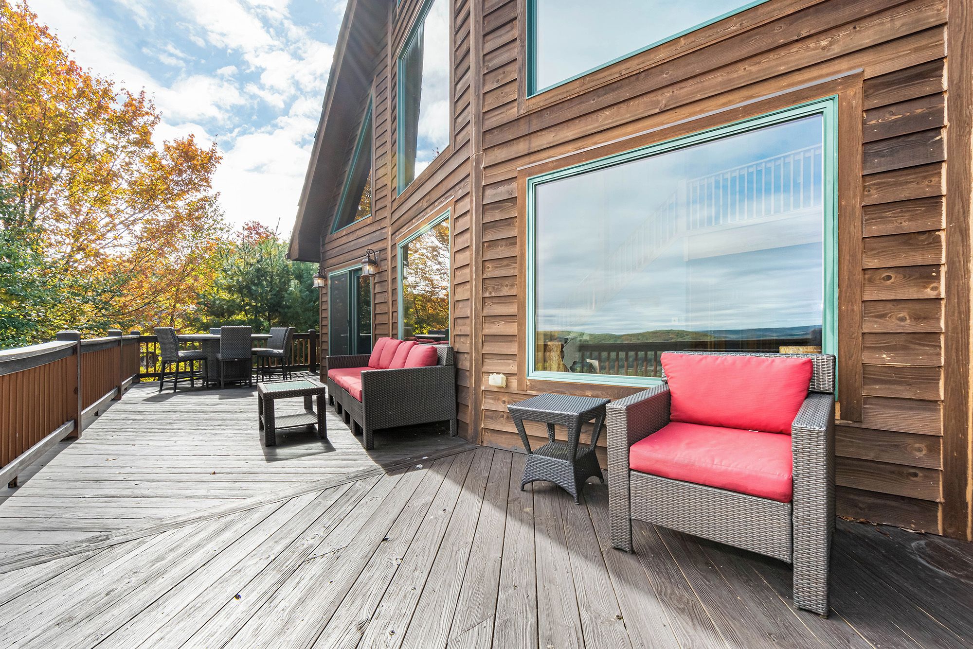A wooden deck with outdoor furniture, including wicker chairs and a sofa with red cushions, overlooking a scenic view.
