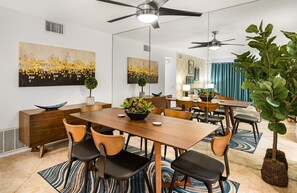A modern dining area with a wooden table, black and wooden chairs, a central plant, and a ceiling fan. - Downtown Oasis. Stylish 2 BR w King beds. Quiet complex w Pool. (Palm Springs)