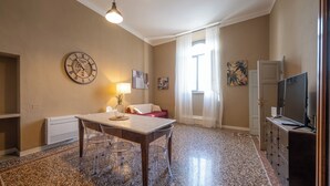 Um quarto espaçoso com uma mesa de jantar, cadeiras, um relógio, uma luminária, um sofá, uma TV e uma janela com cortinas.