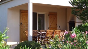 A covered outdoor seating area with wooden chairs and a table, surrounded by flowering plants and a stone fireplace. - Semi-detached mini villa for 6 people in a small, quiet, natural family residence (San-Gavino di Carbini)