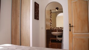 A bedroom with a bed, a wooden door, a bathroom with a sink and mirror, and a wall with a decorative piece. - Semi-detached mini villa for 6 people in a small, quiet, natural family residence (San-Gavino di Carbini)