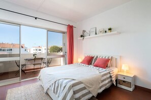 A bedroom with a bed, bedside table, and a window overlooking a cityscape. - Silchoro - Charming, renovated studio with pool and AC (Albufeira)