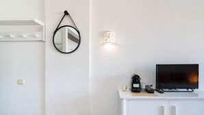 Une chambre moderne avec une coiffeuse blanche, un miroir rond, un luminaire fixé au mur et un téléviseur posé sur une commode blanche.