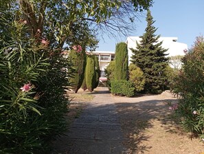 Ein gepflasterter Weg führt zu einem Gebäude mit weißer Fassade, umgeben von Bäumen und Sträuchern.