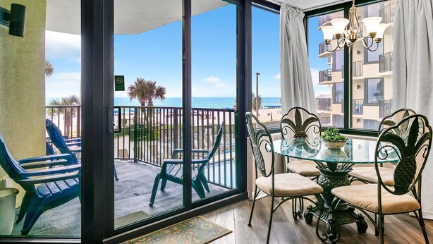 Ein Balkon mit Glastür, der zu einem Poolbereich führt, mit Tisch und Stühlen und Blick auf Strand und Meer.