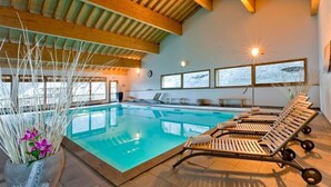 An indoor swimming pool with lounge chairs, wooden beams, and large windows with snow views.