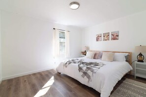 Une chambre à coucher avec un lit, une fenêtre avec des rideaux, une table de chevet avec une lampe et des œuvres d’art accrochées au mur.