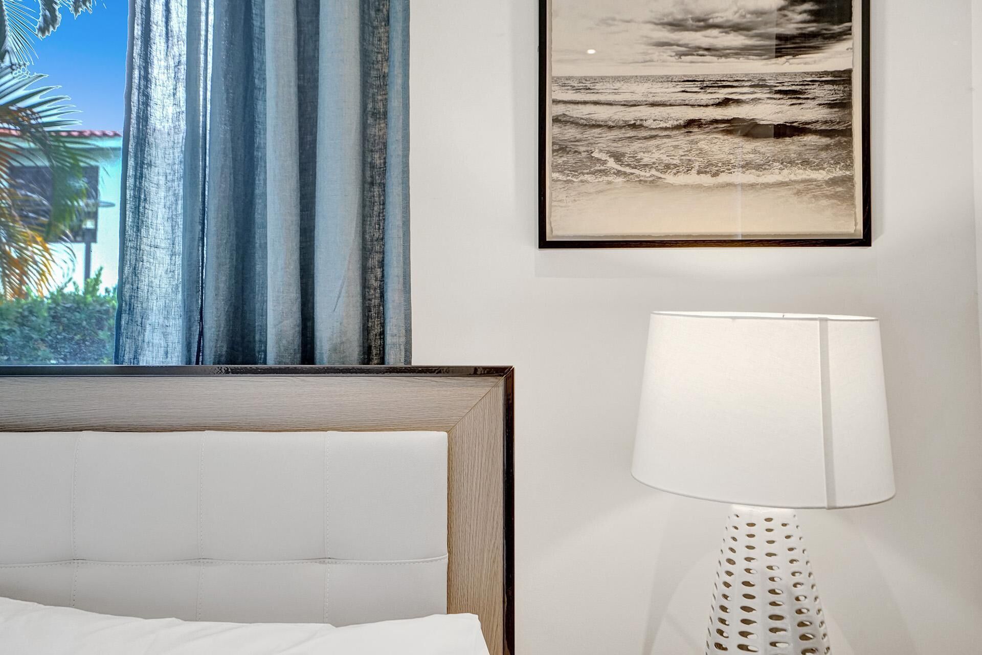 A hotel room with a bed, a window with curtains, a framed picture of the ocean, and a lamp.