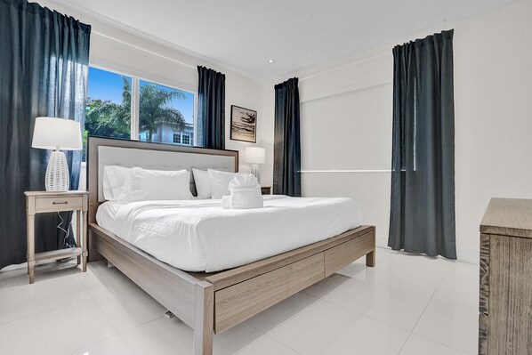 Une chambre d’hôtel moderne avec un lit en bois, du linge de lit blanc, une table de chevet avec une lampe et une vue sur les palmiers par la fenêtre.