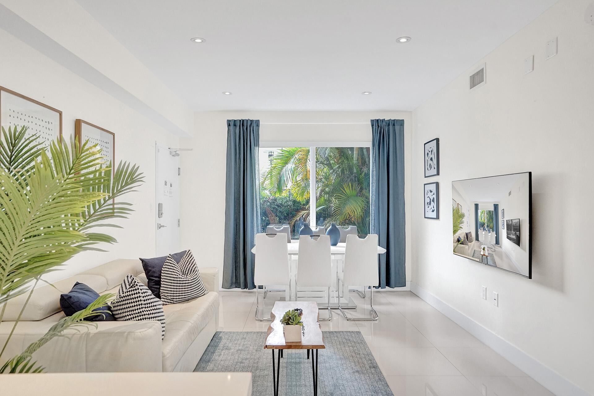 A modern living room with a white sofa, a coffee table, and a dining area with chairs. There are framed pictures on the wall and a large window with curtains.