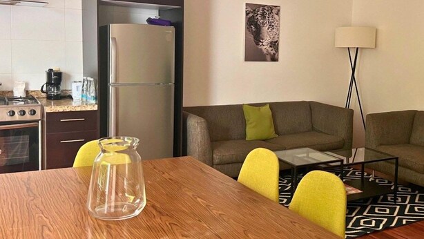 A modern kitchen with a refrigerator, stove, and coffee maker. There is a dining table with chairs and a glass vase. A sofa and a floor lamp are in the living area. - Condesa Residence Amsterdam F (Ciudad de México)