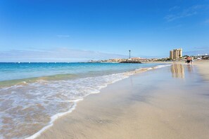 A beach with clear blue water, a sandy shore, and buildings in the background. - Home2Book Dream Apartment Altemar Las Américas (Playa de la Américas)
