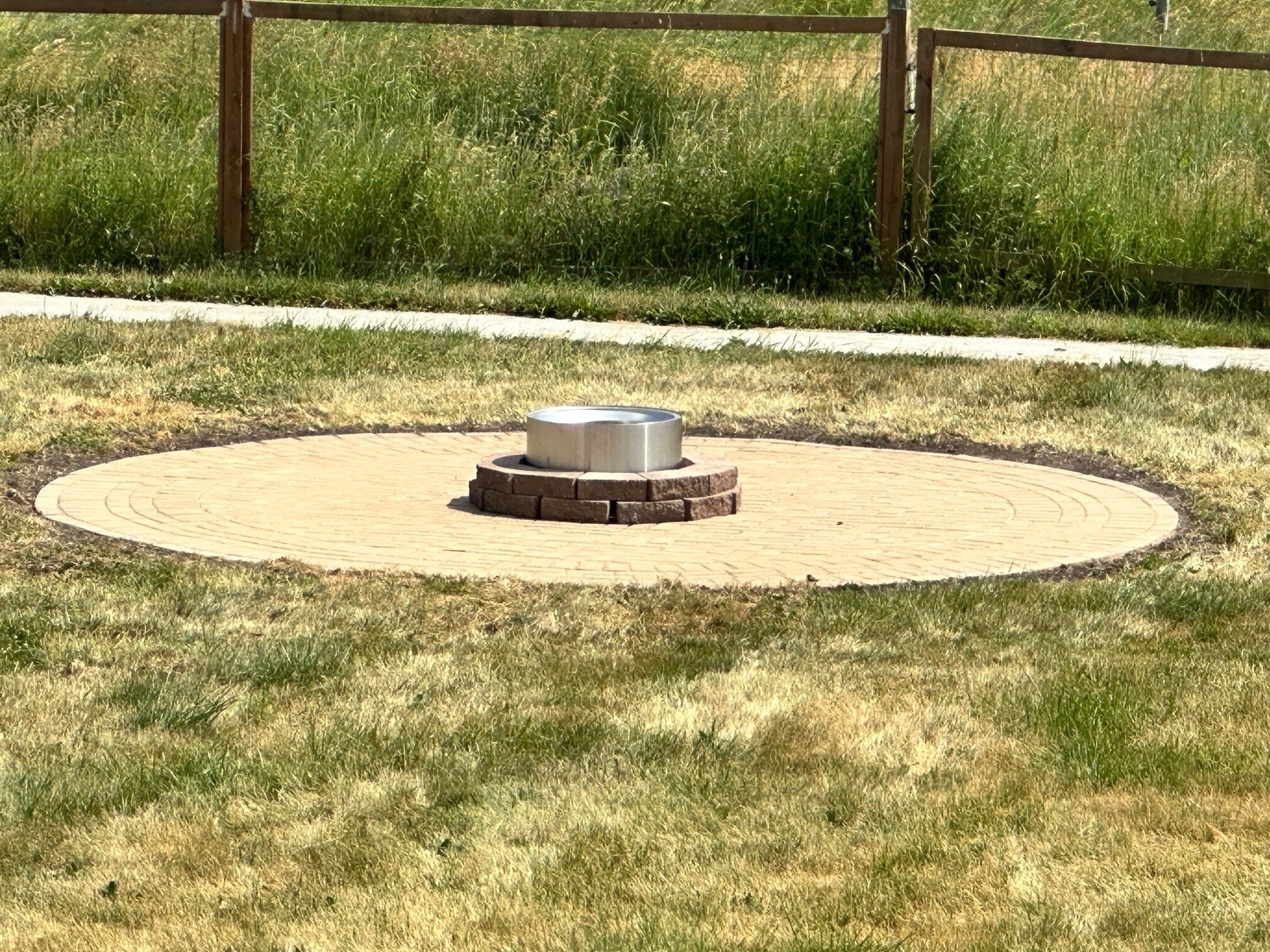 Een rond terrras van baksteen met een centrale metalen constructie, omringd door gras en een houten omheining.