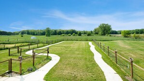 A fenced outdoor area with a white path, surrounded by green fields and trees. - Renovated Dairy Barn – 4 Bedrooms, 5 Beds, 2.5 Baths, Huge, Accessible & Stylish (Byron Center)
