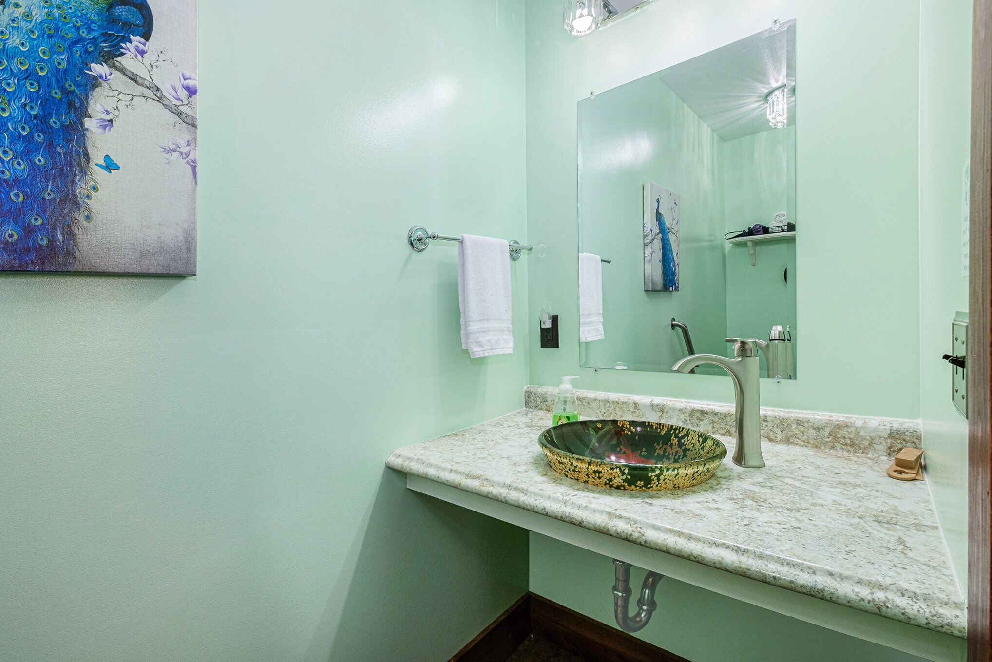 A bathroom with a green wall, a peacock painting, a round sink with a speckled basin, a large mirror, and a towel rack.
