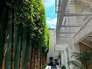 A narrow corridor with vertical bamboo plants on one side and a building with a grid-like ceiling on the other.