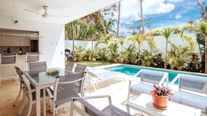 Un espace de restauration extérieur moderne, doté d’une table et de chaises en verre, d’une piscine et offrant une vue sur un jardin luxuriant et le ciel.
