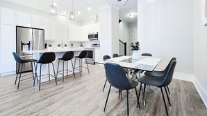 Et moderne køkken med hvide skabe, et rustfrit stål køleskab og en mikrobølgeovn. Der er et spiseområde med et glasbord og sorte stole.