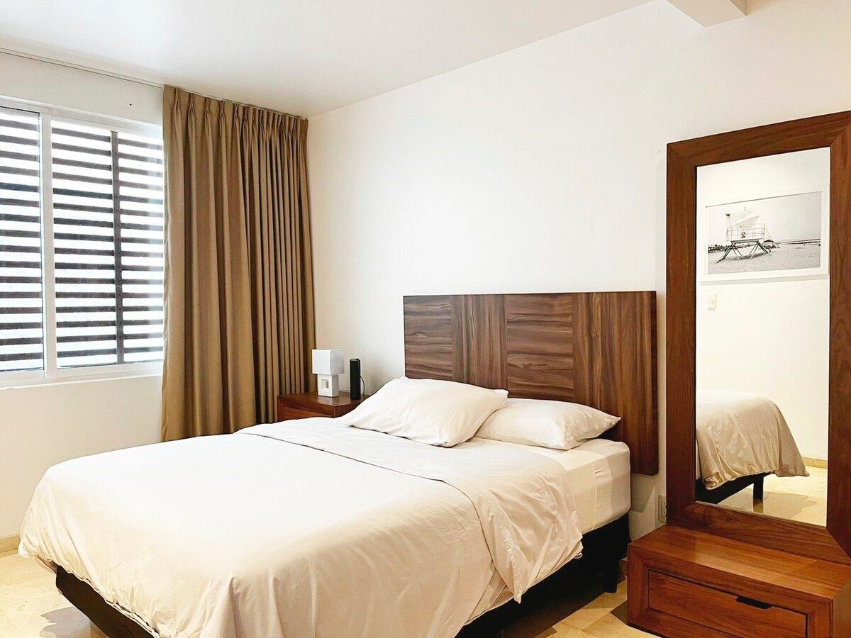 Een hotelkamer met een groot bed, een nachtkastje met een lamp, een houten hoofdbord, een raam met jaloezieën en een spiegel aan de muur.