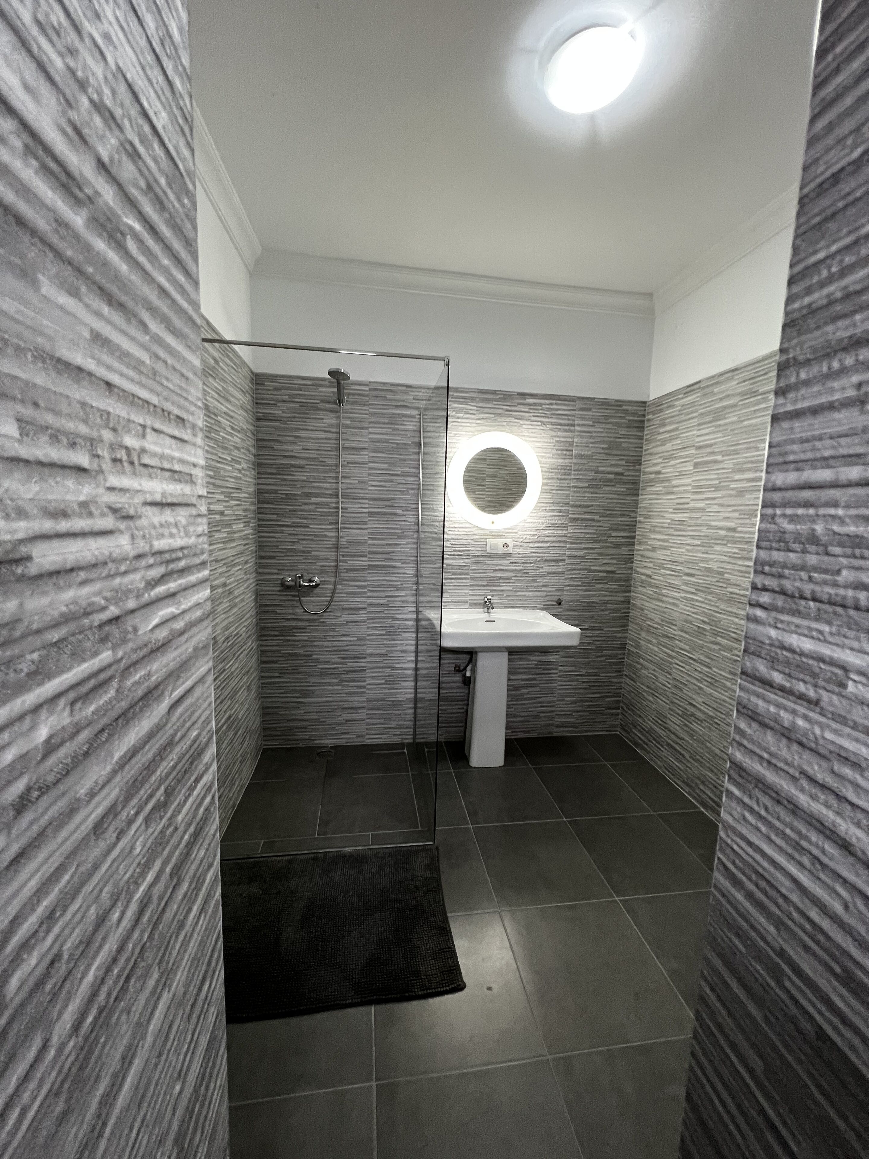 A modern bathroom with a glass shower enclosure, a round mirror, and a pedestal sink.