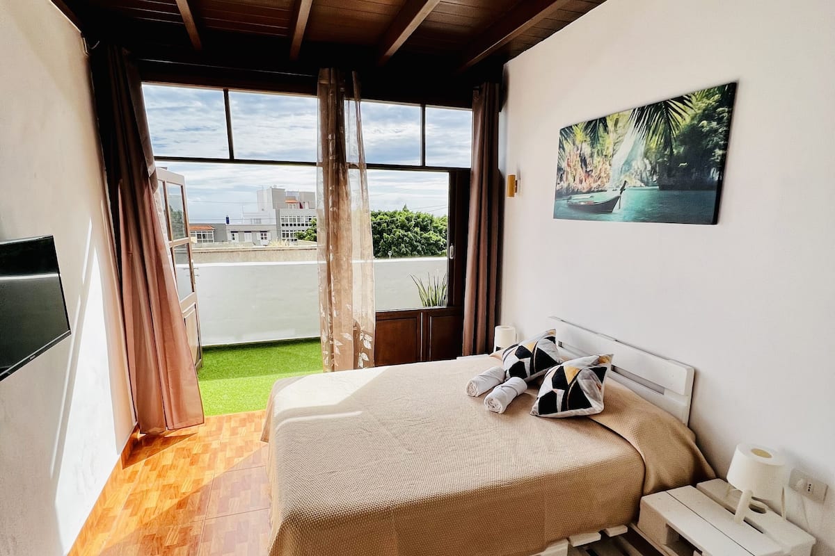 A bedroom with a bed, a nightstand, a painting, and a view of a building and greenery outside.
