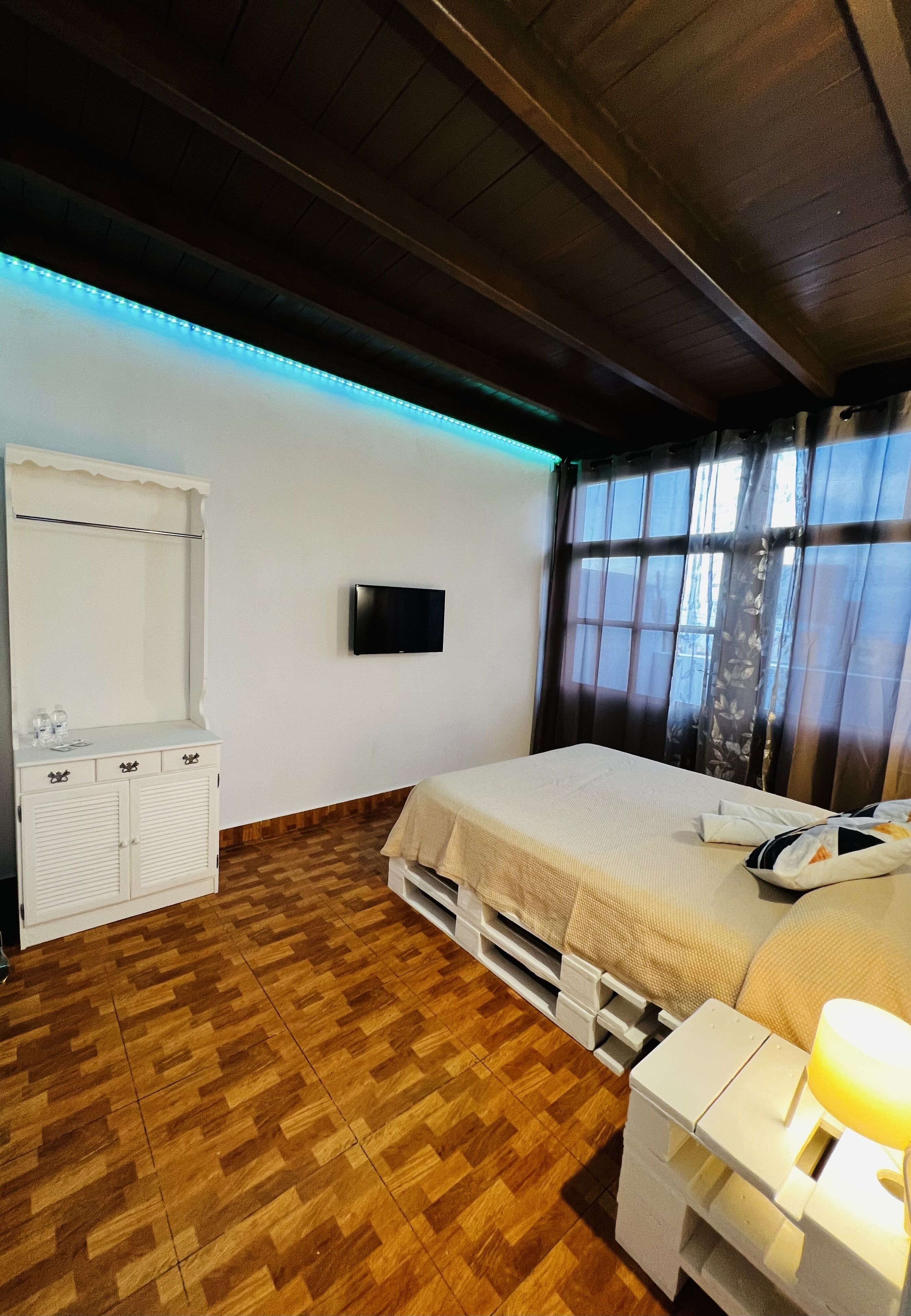 Un dormitorio con suelo de madera, una cama con manta color beige, una mesita de noche con lámpara, un televisor en la pared y un armario.