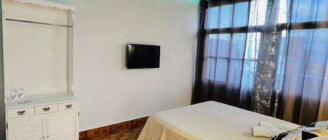Un dormitorio con suelo de madera, una cama con manta color beige, una mesita de noche con lámpara, un televisor en la pared y un armario.