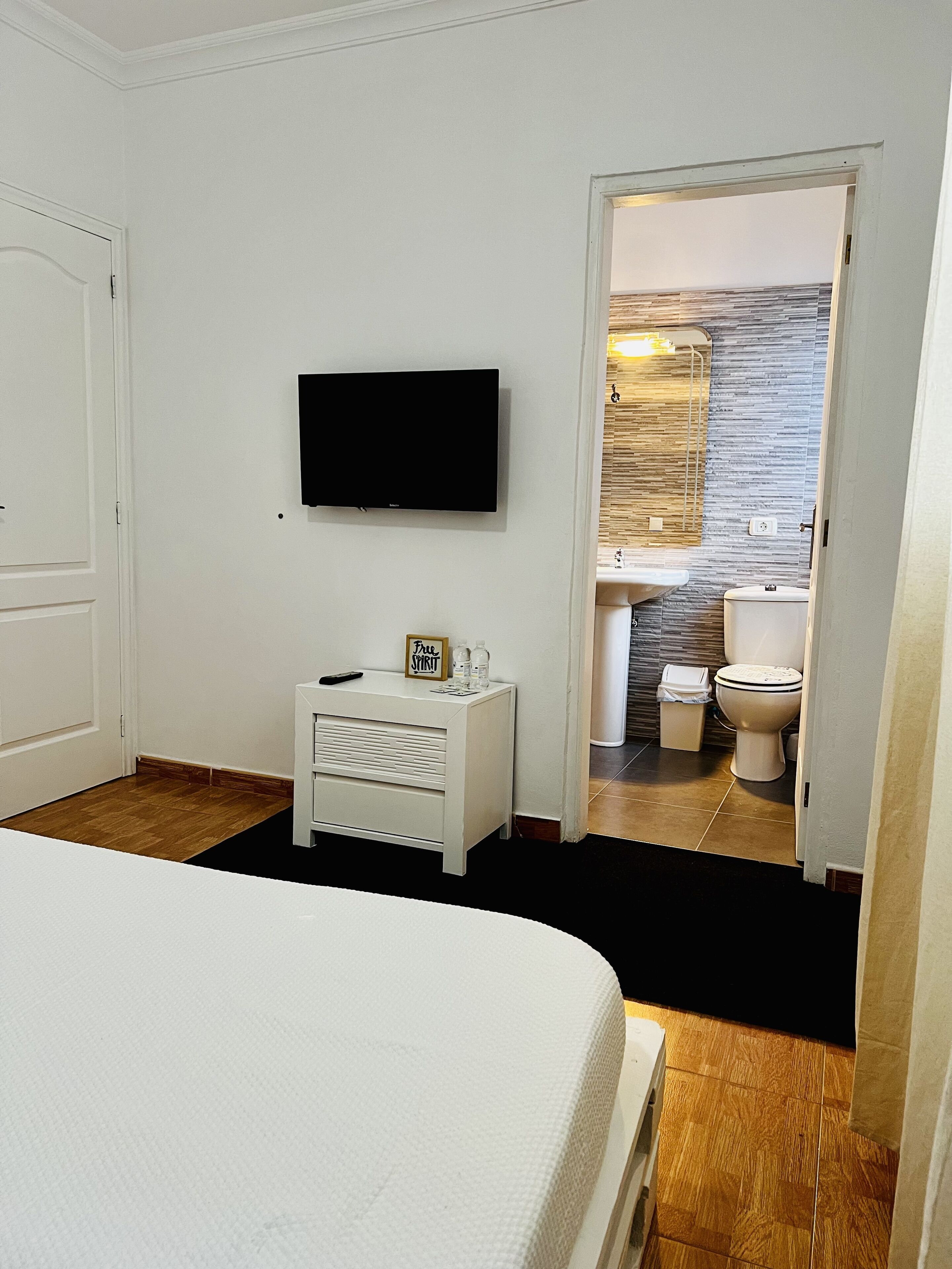 Habitación de hotel con cama, televisor montado en la pared, una mesita de noche pequeña con un control remoto y un cartel que dice 'Free Spirit', y un baño visible a través de una puerta abierta.