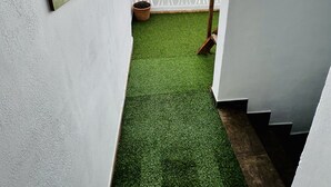 Een smalle gang met groen kunsttapijt, witte wanden en een houten deur die leidt naar een buitenruimte met een witte schutting en een bloempot.