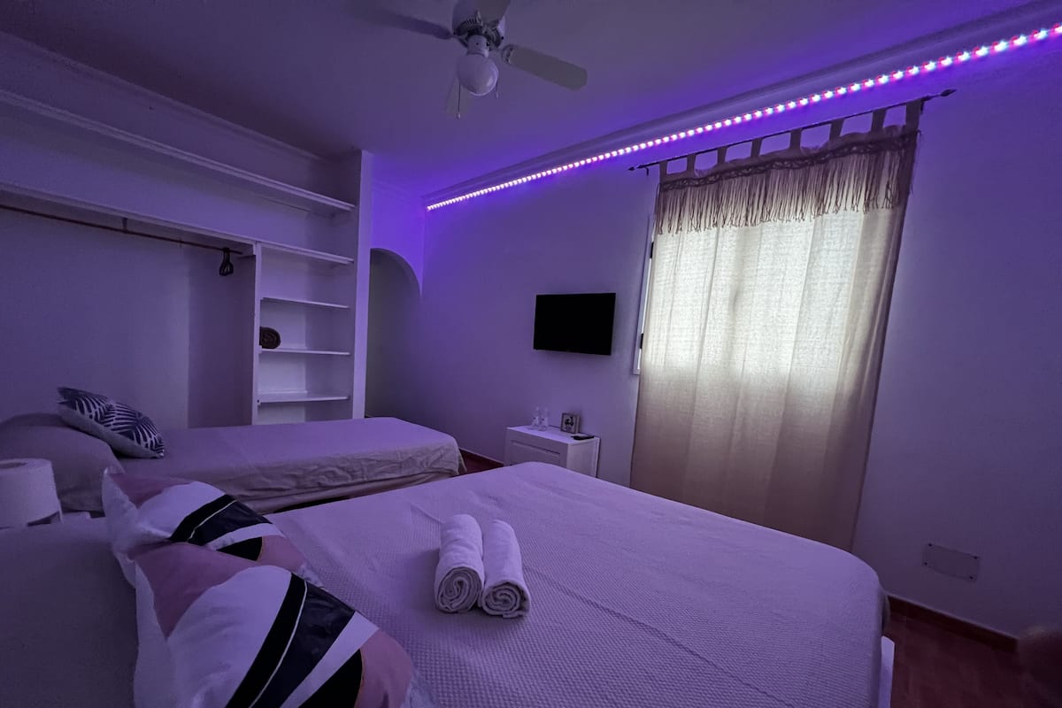 A bedroom with two beds, a ceiling fan, a TV, and LED strip lighting.