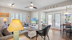 A spacious living room with a sofa, coffee table, and TV, large windows, and a ceiling fan. - 15 SS Cottage - Close to Beach and Village (Siesta Key)