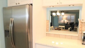 A kitchen with a stainless steel refrigerator, white cabinets, a countertop, and a mirror reflecting a living room with a sofa and a lamp. - 303 Sea Shell- Beautifully Updated Gulfside Condo (Siesta Key)